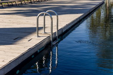 Bu resim, sakin bir su kütlesinin üzerine uzanan ve yüzmek veya rahatlamak için kolay erişim sağlayan parlak metal bir merdiven ile donatılmış ahşap bir rıhtımı gösteriyor..