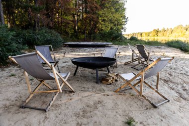 Huzurlu bir kamp ateşi alanı. Zarif katlanır sandalyeler, yakındaki yakacak odunları ile süslü bir ateş çukurunun etrafına dizilmiş, yemyeşil ağaçlar ve dingin sulara karşı kurulmuş..