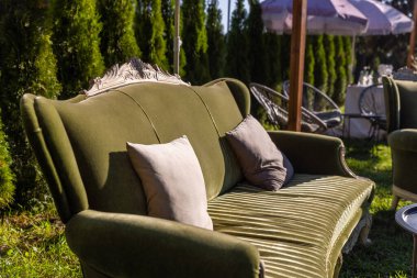 Yumuşak yastıklı davetkar yeşil bir kanepe yeşillik ve açık hava mobilyalarının sergilendiği güzel bir bahçe arkaplanına zarafet katar, rahatlama ve rahatlama aktiviteleri için mükemmeldir..