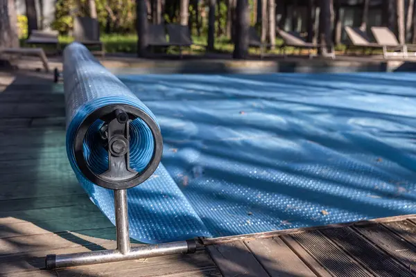 Bu resim, ağaçlar ve şezlonglarla zenginleştirilmiş sakin bir ortamda, ahşap bir güvertede duran zarif mavi kapaklı bir havuz örtüsü silindirini tasvir ediyor. Rahatlamak için mükemmel..