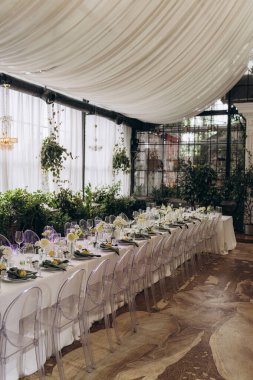 Çiçekler, temiz sandalyeler ve zarif sofra takımlarıyla süslenmiş uzun bir masa. Yeşillik ve örtülü tavan toplanma için davetkar bir atmosfer yaratıyor..