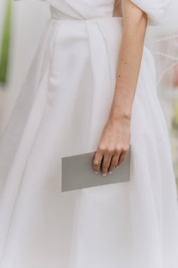 Görüntüde zarif bir el ışıl ışıl bir yüzükle süslenmiş, zarif gri bir zarf tutan, zarif beyaz bir elbiseyi mükemmel bir şekilde tamamlayan, zarif bir el sergileniyor..