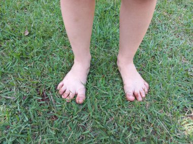 Taze yeşil çimlerin üzerinde duran bir çocuğun çıplak ayaklarının yakın plan görüntüsü, çocukluğun masumiyetini ve doğayla olan bağlantısını gösteriyor. Ayak parmakları açık havada oynadığı için biraz kirlidir. Kaygısız bir yaz anını yakalar..