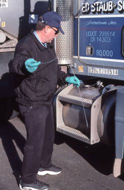 Government agent checks truck fuel tank for red dyed diesel. clipart