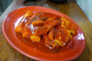Çiğnenmiş kırmızı soslu pirinç kekleri ve susam tohumları ve yeşil soğanla kaplı baharatlı Kore tteokbokki. Bağımlılık yaratan tadı ve tatmin edici dokusuyla bilinen popüler bir sokak yemeği..