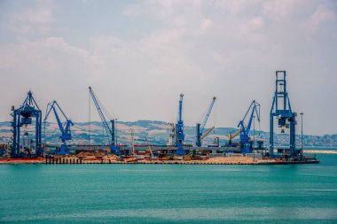 İtalya 'nın Ancona kentindeki hareketli bir kargo limanının panoramik görüntüsü, kıyı tepeleri ve turkuaz sularına karşı yükselen mavi turnalar.