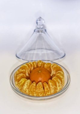 An unpeeled orange rests at the centre of a ring of peeled wedges beneath a glass dome, creating a bright citrus focus and a fresh, inviting arrangement