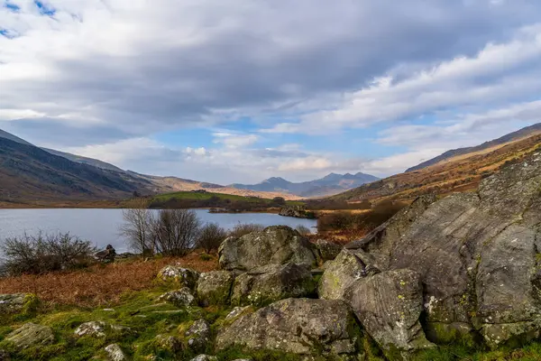 Framed by winter branches and Large Boulders, Llyn Gwynant showcases Snowdonias rugged peaks, offering a quintessential Welsh mountain panorama