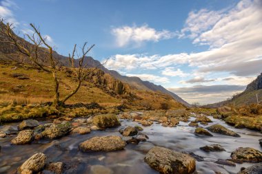 Silky river weaving through boulderstrewn Snowdon foothills valley beneath drifting cumulus clouds at golden hour