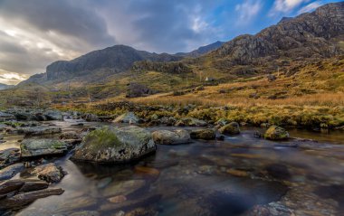 Longexposure watercourse gliding over mossclad boulders through Snowdonia valley girded by rugged peaks under sweeping clouds