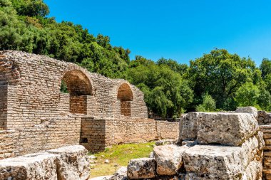 Akdeniz ormanları ve açık bir yaz gökyüzü tarafından çerçevelenmiş Butrint 'te yıpranmış kireçtaşı blokların üzerinde yükselen ikiz Roma varil kemerleri