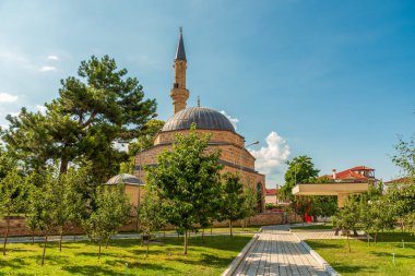 Arnavutluk 'un Korca kentindeki ağaçlarla kaplı bir park alanının üzerinden yükselen ince minare ve kubbeli Mirahori Camii bulutlu yaz gökyüzünün altında