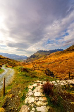 Çakıl taşlı patika, kenevir kaplı kıyıyı geçip Pen-y-Geçidi 'ne doğru dönüyor ve Snowdon Dağı' nın heybetli yamaçları dinamik bir gökyüzü altında.