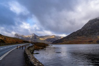 Llyn Ogwen 'in yanındaki kıvrımlı göl yolu dalgalı yansımalarla kara kara kara kara kara bulutların altında engebeli tepelere çıkıyor.