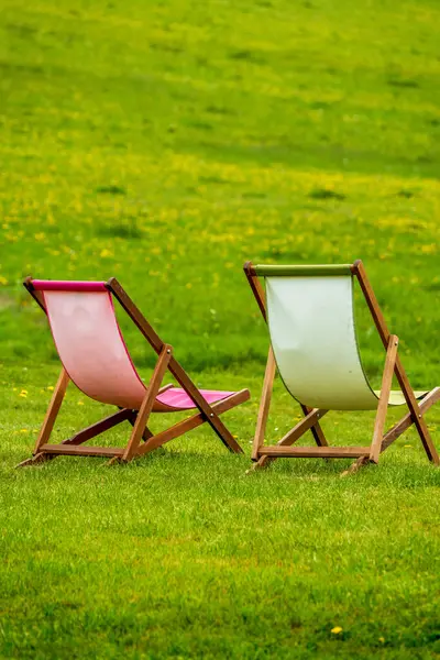 Pastel brandalı iki ahşap iskemle yayılmış gün ışığı altında yeşil çimenli bir yamaçta dizilmiş.