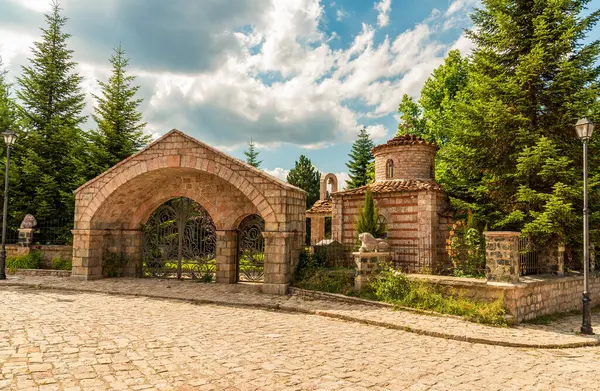 Arnavutluk 'un Voskopoje kentindeki kaldırımlı bir plazada demirden kapılar ve çam siluetleriyle çerçevelenmiş taş kemerli kapı ve bitişik kubbeli pavyon