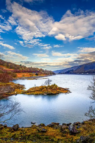 Rüzgârlı meşe ve kayalık kıyılarla çevrili buzullu tarn, kayan bulutların altında, Snowdon Dağı eteklerinde.