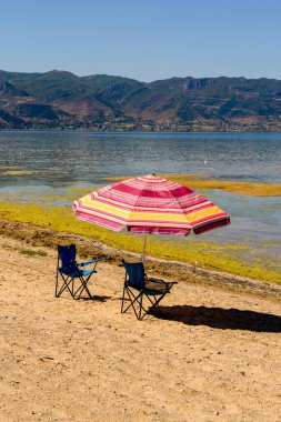 Arnavutluk 'un Ohri Gölü, Pogradec kentinde temiz su ve dağ manzaralı kumlu bir göldeki renkli şemsiye ve katlanan sandalyeler