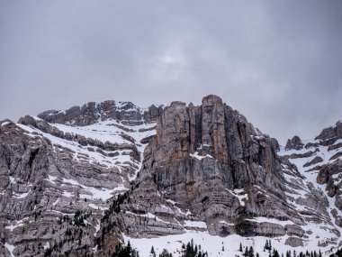 İspanyol Pireneleri 'ndeki engebeli Roca Verda kaya yüzeyinin geniş bir görüntüsü, dramatik, bulutlu bir gökyüzünün altında, dağınık kozalaklı ağaçlarla korunmasız kireç taşı ve kar parçaları sergiliyor..