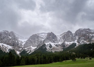Pireneler 'deki engebeli Sierra del Cadi' nin Prat del Cad 'den çekilmiş geniş bir görüntüsü. Huysuz, bulutlu bir gökyüzünün altında, yeşil bir çayırın üzerinde dramatik uçurumlar yükselir..