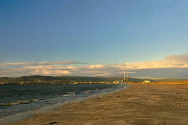Açık mavi gökyüzü ve birkaç bulutu olan bir sahil. Kumsal boş ve su sakin.