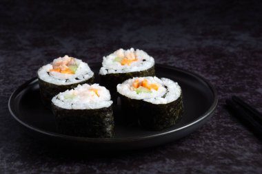 Siyah bir tabakta dört suşi rulosu. Ekmekler yosun ve pirinçten yapılmıştır. Tabak karanlık bir yüzeye yerleştirilmiş.