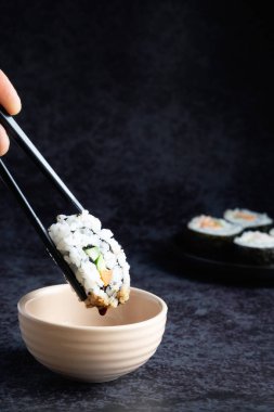 Yemek çubuklarının yakın plan çekimlerinde bir sos kabının üzerinde suşi rulosu tutuyorlar. Arka planda diğer suşi parçaları bulanık.