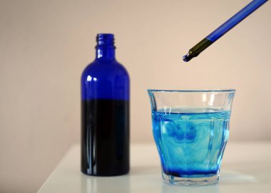 Görüntü, su ve metilen mavisi bardağın yanında metilen mavisiyle dolu bir şişe ve camın üzerinde bir pipet gösteriyor..