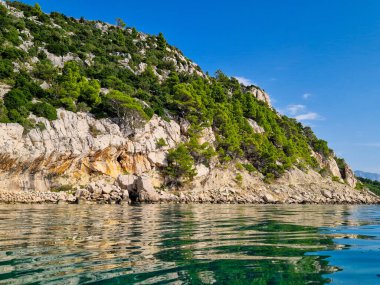 Hırvatistan 'daki Adriyatik Denizi' ndeki mavi gökyüzünü yansıtan berrak ve sakin sulara sahip kayalık bir kıyı şeridi manzarası