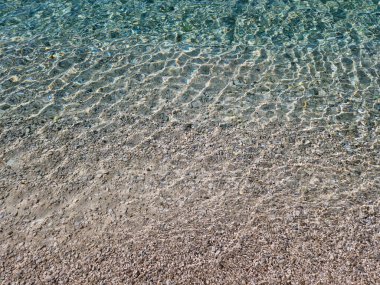 Çakıl taşlı bir plajın üzerinde şeffaf deniz suyu. Güneş ışığı yansımalarıyla parıldıyor.