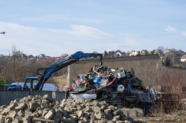 Hidrolik bir kazıcı, arka planda köyü olan bir geri dönüşüm sahasında hurda metal organize ediyor.