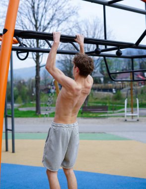 Atletik genç adam dışarıda egzersiz yapıyor, metal fitness çubuğunda barfiks çekiyor.