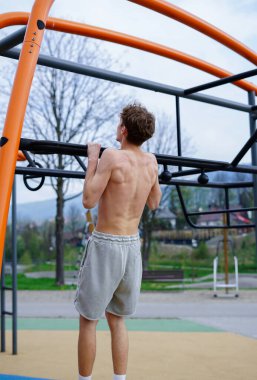 Atletik genç adam dışarıda egzersiz yapıyor, metal fitness çubuğunda barfiks çekiyor.