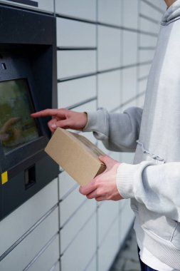 Gri kapüşonlu erkek, karton pakette parsel dolap istasyonunun dokunmatik ekran terminalini kullanıyor.