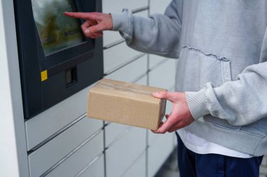 Gri kapüşonlu erkek, parsel dolap istasyonunun dokunmatik ekran terminalini kullanarak karton paket topluyor..
