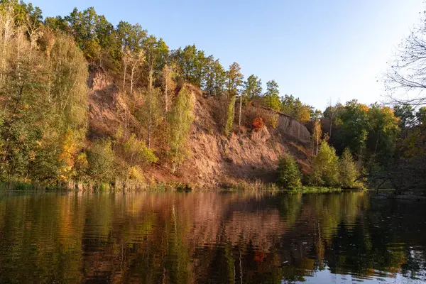 Batan güneşin kırmızısındaki nehir yamacındaki renkli sonbahar ağaçları.