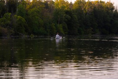 Yalnız bir kanocu beyaz bir kanoda kürek çeker ve sık bir ormanın gölgesinde suyun üzerinde dinlenir.