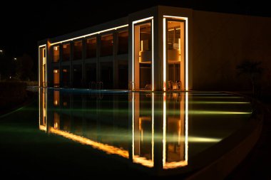 Lüks otelin aydınlanmış modern mimarisi geceleri huzurlu havuza yansıyor.