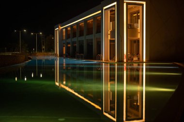 Lüks otelin aydınlanmış modern mimarisi geceleri huzurlu havuza yansıyor.