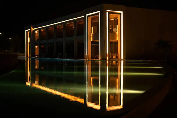 Lüks otelin aydınlanmış modern mimarisi geceleri huzurlu havuza yansıyor.