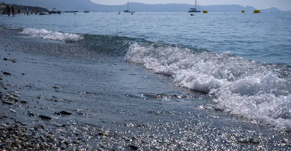 a foamy wave rolls onto a pebble sea beach with boats in the distance