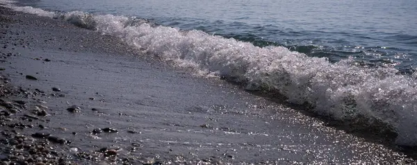 a straight foamy wave rolls onto a pebble sea beach in the rays of the sun