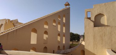 Çan kulesi ve kemerli pencereleri olan bir bina. Bina ten renginde. Binanın çatısında dikilen insanlar var. Jaipur, Hindistan 'da Jantar Mantar