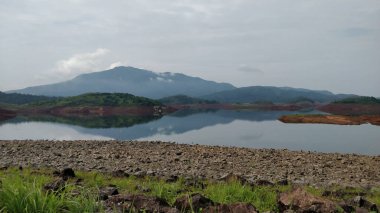 Bir dağ sırası bir gölün suyuna yansıtılır. Gökyüzü bulutlu ve arka planda dağlar var. Banasura Sagar Barajı, Kerala, Hindistan