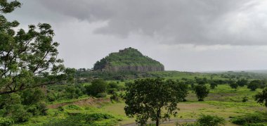Bir dağ yemyeşil bir tarlanın arka planındadır. Gökyüzü bulutlu ve atmosfer huzurlu. Kailasa Tapınağı, Ellora Mağaraları, Hindistan