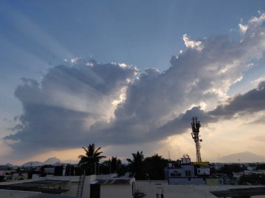 Scenic urban sunset with dramatic sun rays piercing through clouds, silhouetting palm trees, rooftops, and a telecommunications tower against a distant mountain backdrop. clipart