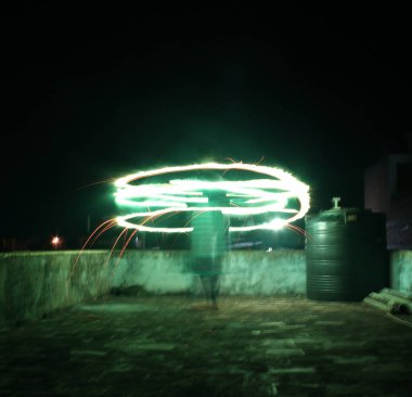 Night photography of a person spinning a green glowing light, creating circular trails in the dark sky. Captured with long exposure, featuring a rooftop setting and vivid motion effects. clipart