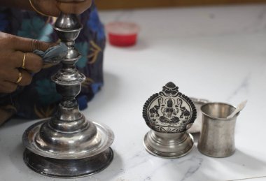 Close-up of a person polishing a silver lamp, with traditional Indian silverware, including a Lakshmi idol and a small cup, on a marble surface. Represents culture, tradition, and devotion. clipart