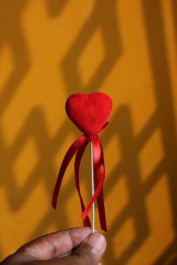 Minimalist photo of a red velvet heart on a stick with ribbons, held against a warm yellow wall with geometric shadows. Perfect for Valentine's Day, love themes, and creative concepts. clipart