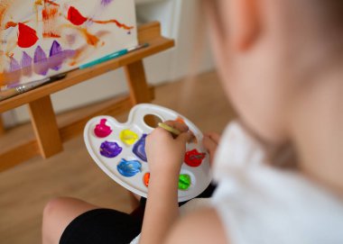 Bir çocuğun ellerinin, tuval üzerinde çalışması için fırçayla bir paletten boya alırken yakın plan fotoğrafı..